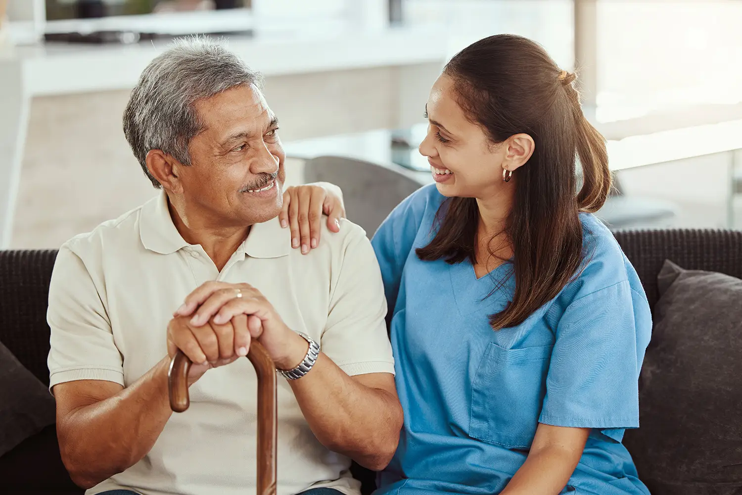 An elderly man with gray hair, wearing a beige polo, smiles while holding a cane. A woman in blue scrubs sits beside him, smiling and resting her hand on his shoulder. They are sitting in a well-lit room, benefiting from the excellent in-home hospice care that Phoenix, AZ offers for comfort and warmth.